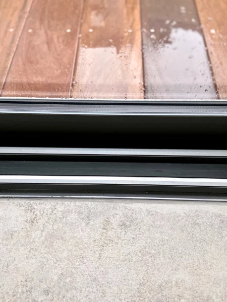 Gros plan du passage de l'intérieur vers l'extérieur de la maison. Rail de porte-fenêtre coulissante, sol en béton ciré et bois sous la pluie côté terrasse