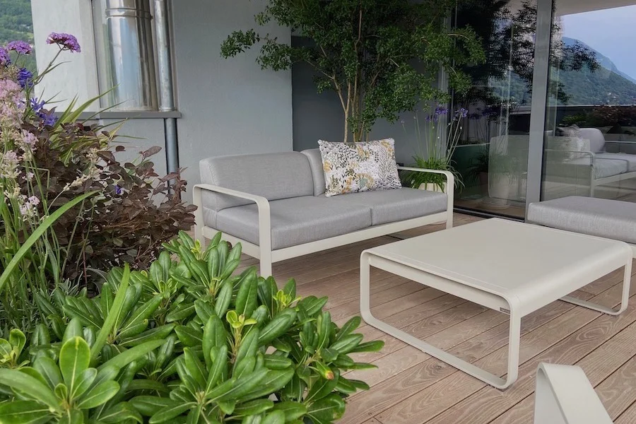 Un coin de balcon aménagé avec un canapé d'extérieur en métal et coussins tissu gris avec un coussin fleuri et une table basse assortie devant. Au premier plan une jardinière avec de la verveine de Buenos Aires et un Pittosporum de Chine