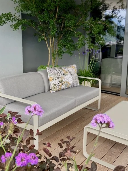 Un coin de balcon aménagé avec un canapé d'extérieur en métal et coussins tissu gris avec un coussin fleuri et une table basse assortie devant